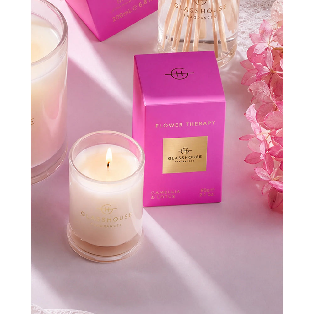 A lit white candle in a clear glass jar sits beside its bright pink box labeled 'FLOWER THERAPY' and 'GLASGOWHOUSE FRAGRANCES.' A matching reed diffuser bottle is partially visible behind it, with pink flowers and a soft pink surface in the background.