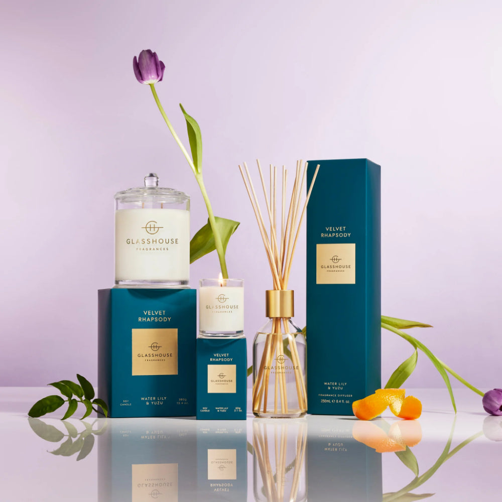 Glasshouse Fragrances products including candles and diffuser on a reflective surface with a light purple background.