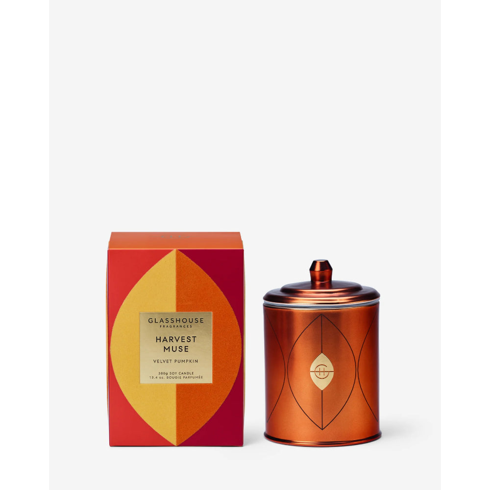 A GLASSHOUSE FANTASY 'Harvest Muse' candle shown with its red and yellow geometric packaging box and a metallic copper-colored tin container with a lid.