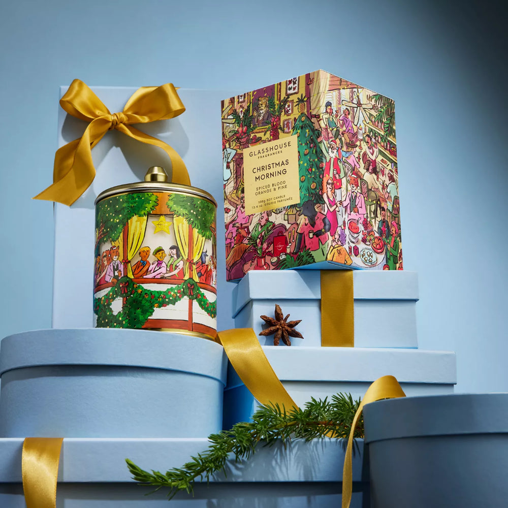 A styled product display featuring a round tin and a tall rectangular box from Classique Candle Co., both decorated with festive Christmas illustrations and tied with gold ribbons, set against a light blue background.