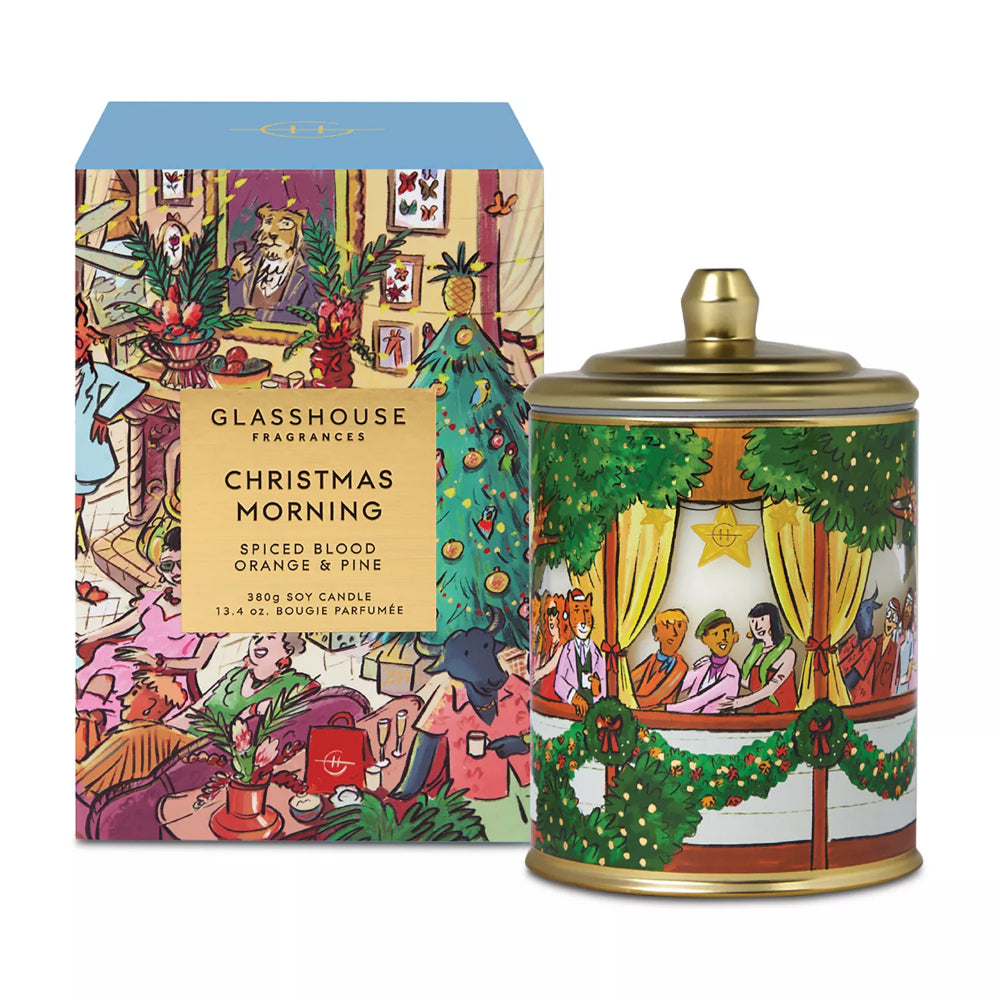 A Christmas-themed scented candle by Glasshouse Fragrances, shown with its illustrated retail box and the matching metal tin candle. The packaging features a busy holiday illustration with people, ornaments, and a tree, while the candle tin has a green garland and gold-toned lid.