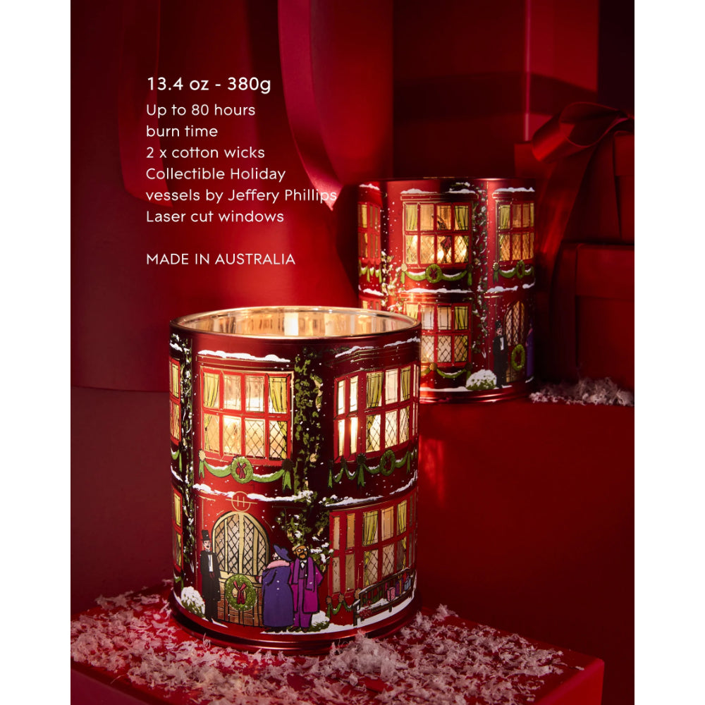 Two Jeffrey Phillips collectible holiday candle holders shaped like red London tube stations, decorated with green ivy and snow, lit with warm yellow flames. The image features promotional text on the left and is set against a dark red background with festive red ribbons and faux snow.