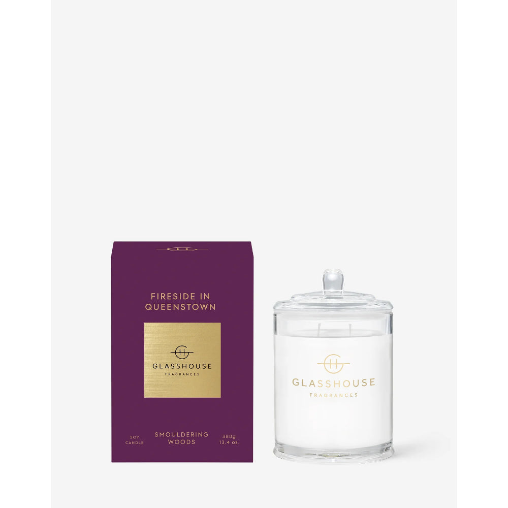 A clear glass jar candle with a white wax fill and lid, shown beside its deep purple box. The packaging features a gold label with the brand name 'GLASSHOUSE FRAGRANCES' and the scent name 'FIRESIDE IN QUEENSTOWN'.