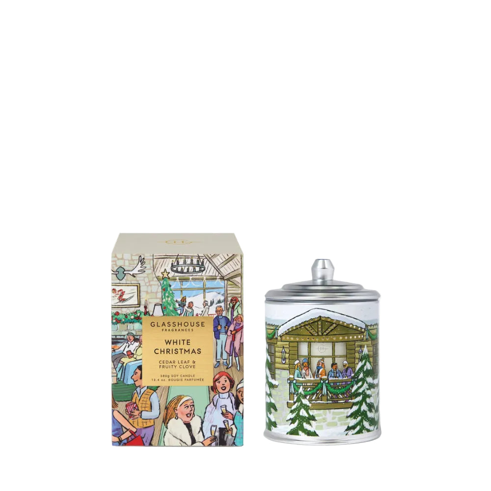 A Christmas-themed scented candle and its matching retail box by Glasshouse Fragrances. The candle is a silver glass jar with a lid, decorated with a festive illustration of a log cabin, people by a fireplace, and evergreen trees in white and green.