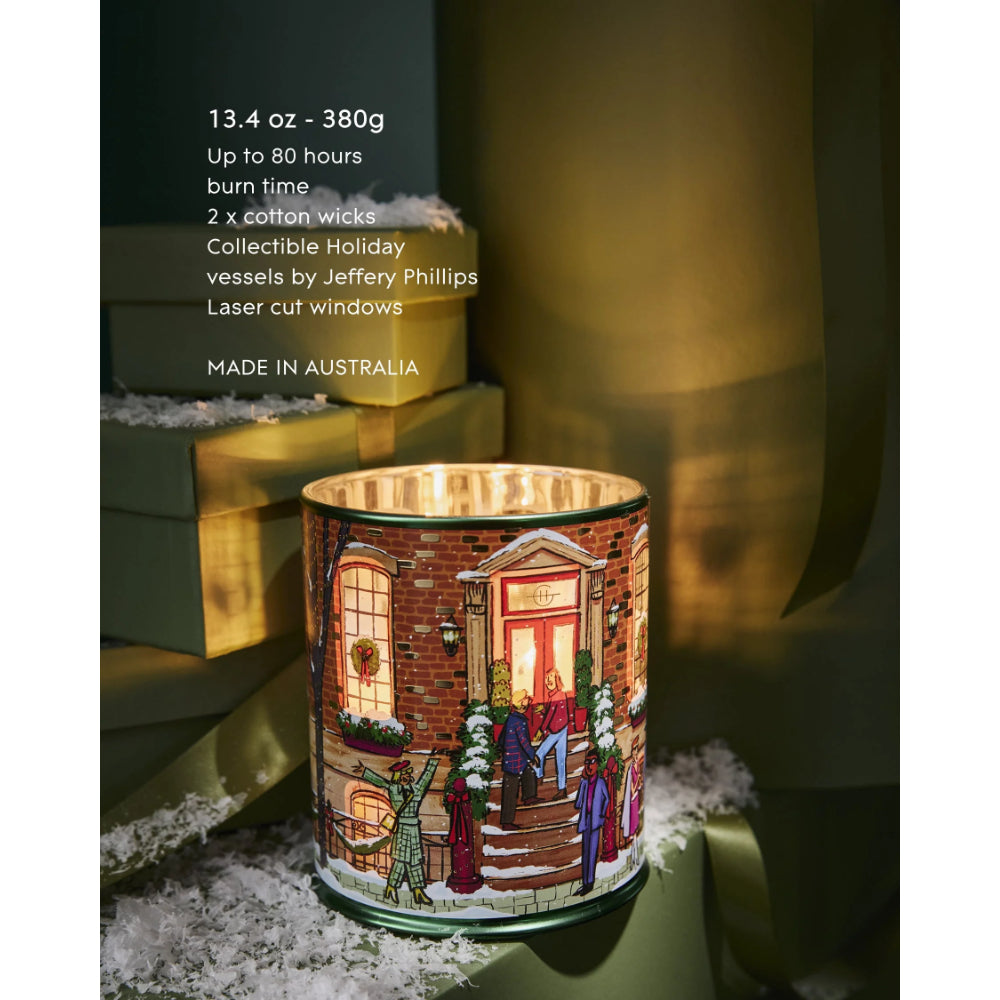 A lit collectible holiday candle in a cylindrical glass vessel printed with a winter village scene, set on a dark surface with stacked green boxes and ribbon in the background. White text on the left lists product details 