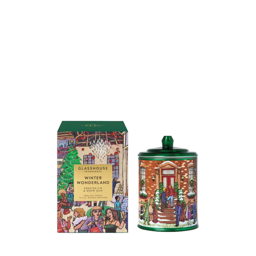 A Christmas-themed scented candle and its matching box from the brand GLASSHOUSE PARADISE, featuring a colorful winter village illustration on both the candle container and the packaging.