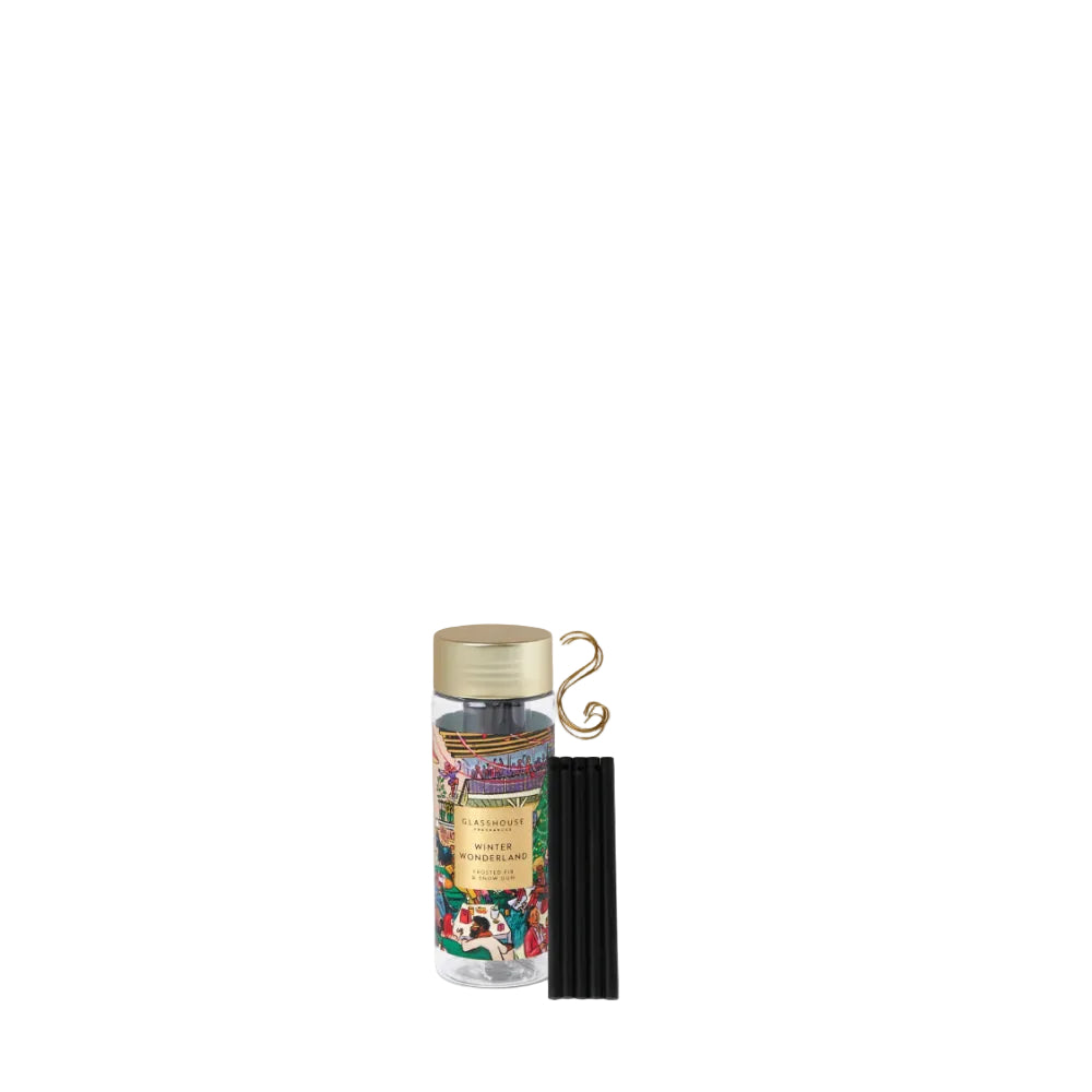 A small clear bottle with a gold cap and a colorful illustrated label, shown next to a black reed diffuser stick and a gold decorative loop, on a plain white background.