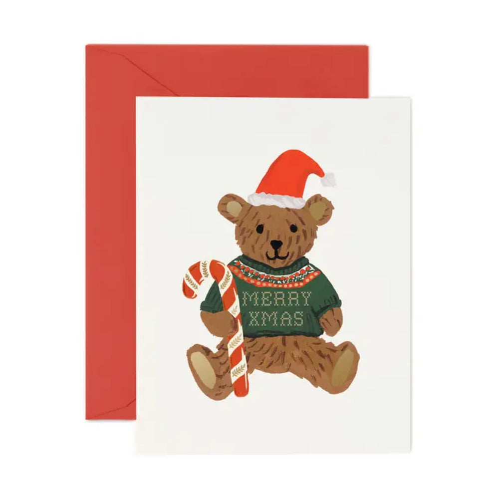 A Christmas greeting card featuring a brown teddy bear wearing a red Santa hat and a green sweater that says 'MERRY XMAS', shown with a red envelope behind it on a white background.