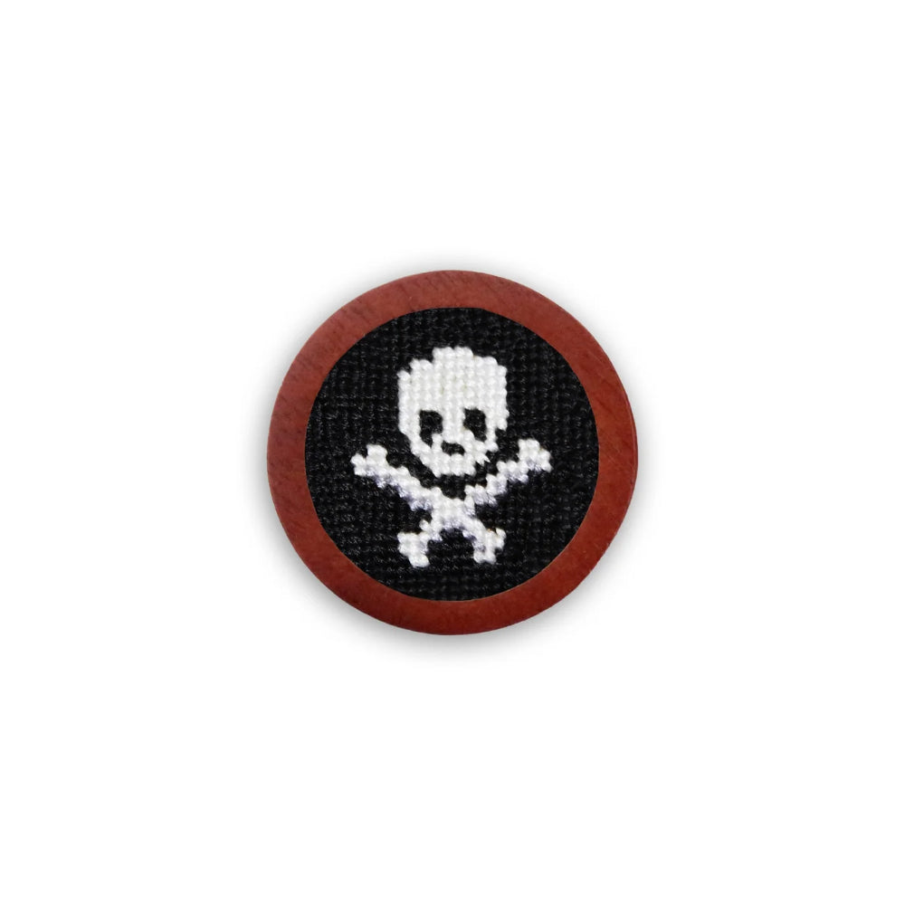 A circular wooden golf ball marker with a needlepoint patch featuring a white stitched skull and crossbones design on a black background, surrounded by a natural brown wood border.
