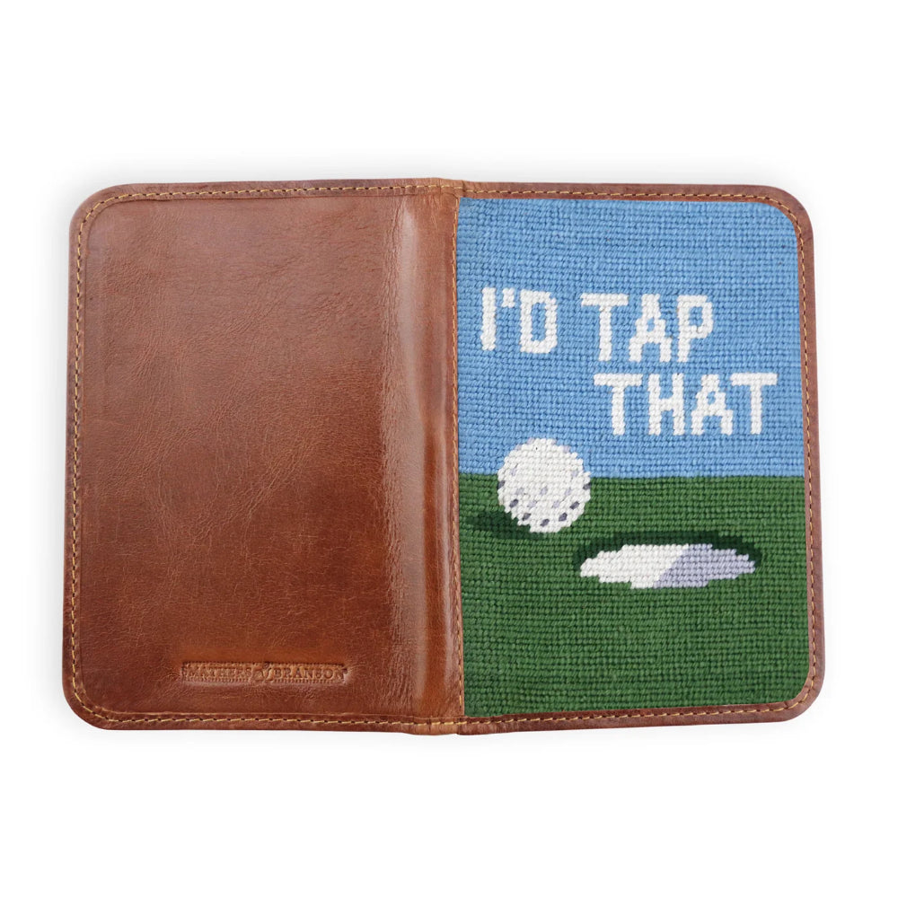 A brown leather bifold gold scorecard holder shown open, featuring a colorful needlepoint panel on the right side with a blue sky, green grass, a white golf ball, and the text 'I'D TAP THAT'.