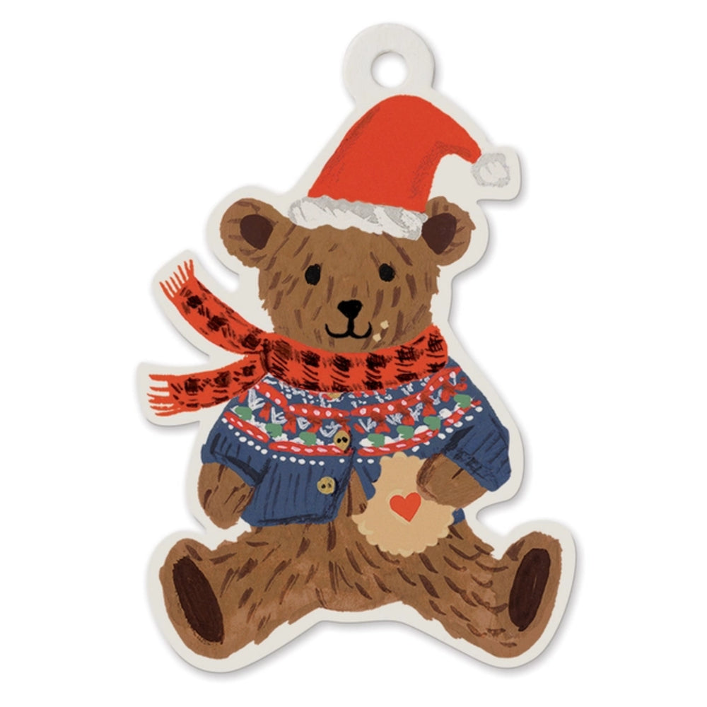 A die-cut teddy bear ornament or sticker featuring a brown bear wearing a red Santa hat with white trim, a red patterned scarf, and a blue sweater with colorful accents, holding a beige heart with a small red heart on it.