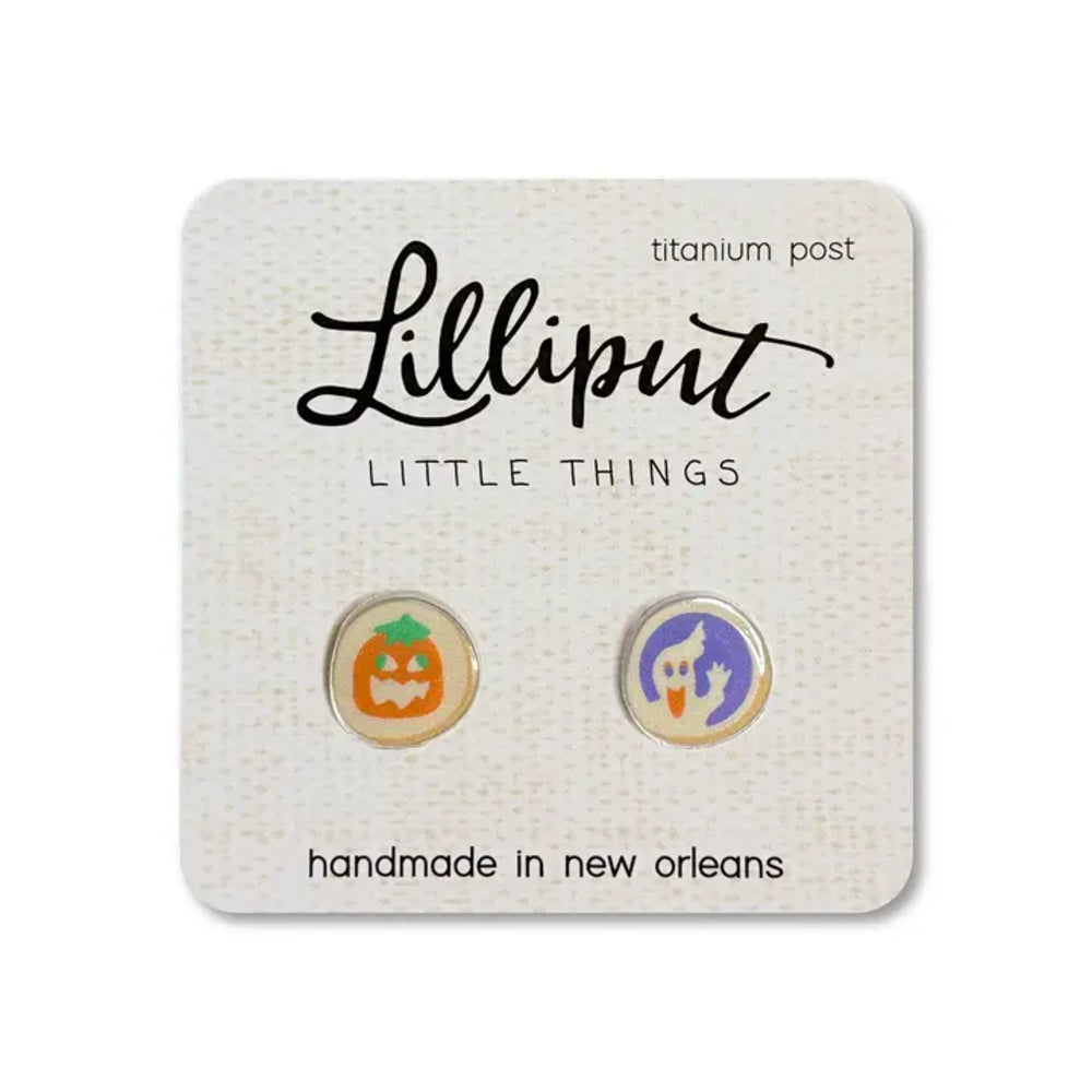 A pair of Lilliput Little Things enamel stud earrings on a textured white display card. The left earring features an orange pumpkin face, and the right earring features a purple ghost with a white face, both set on titanium post backs.