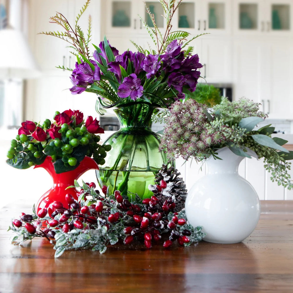 Decorative arrangement of flowers in a green glass vase with a wreath on a wooden surface with a blurred kitchen background.