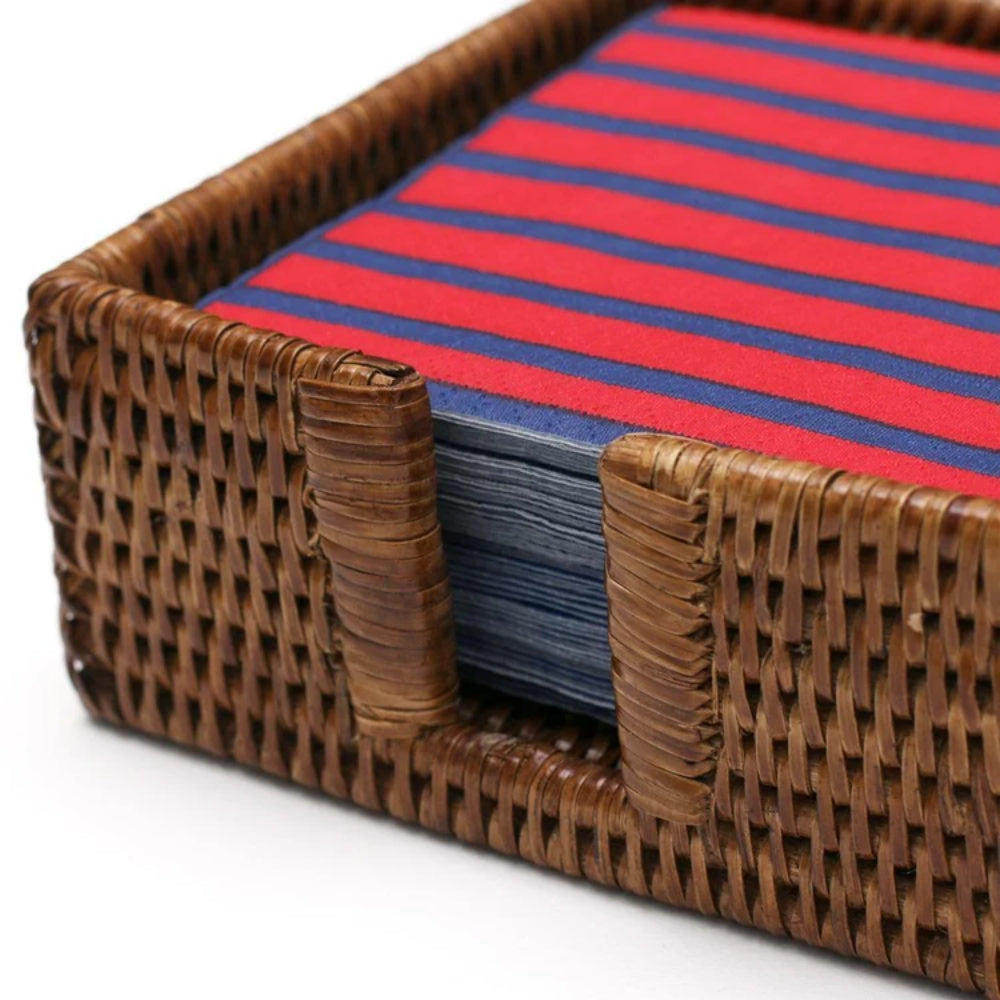 A close-up of a brown woven wicker napkin holder holding a stack of folded red and navy striped fabric pieces, photographed on a white background.