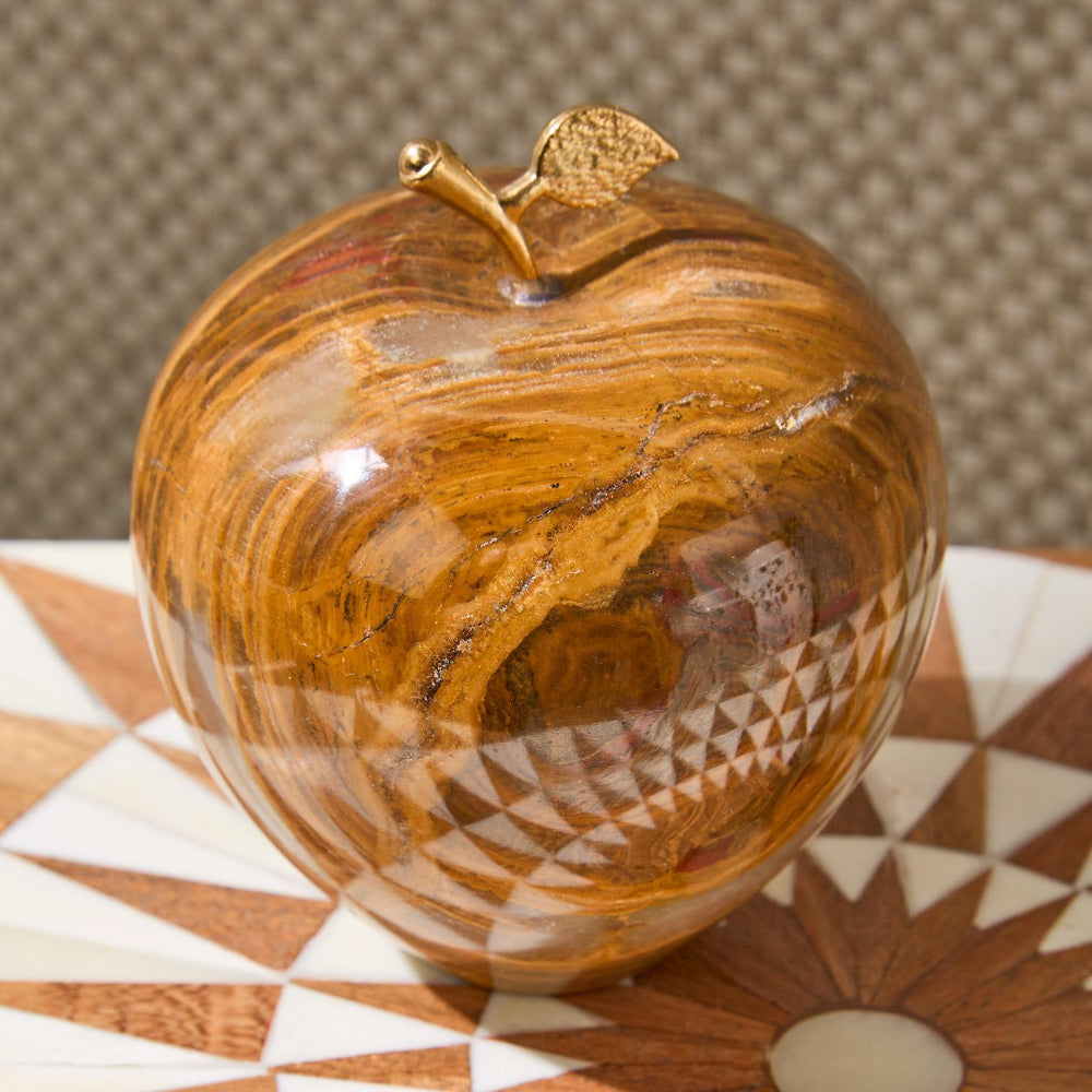 A decorative apple-shaped object with a glossy golden-brown marbled finish and a small metallic gold stem and leaf, shown on a patterned surface against a textured neutral background.