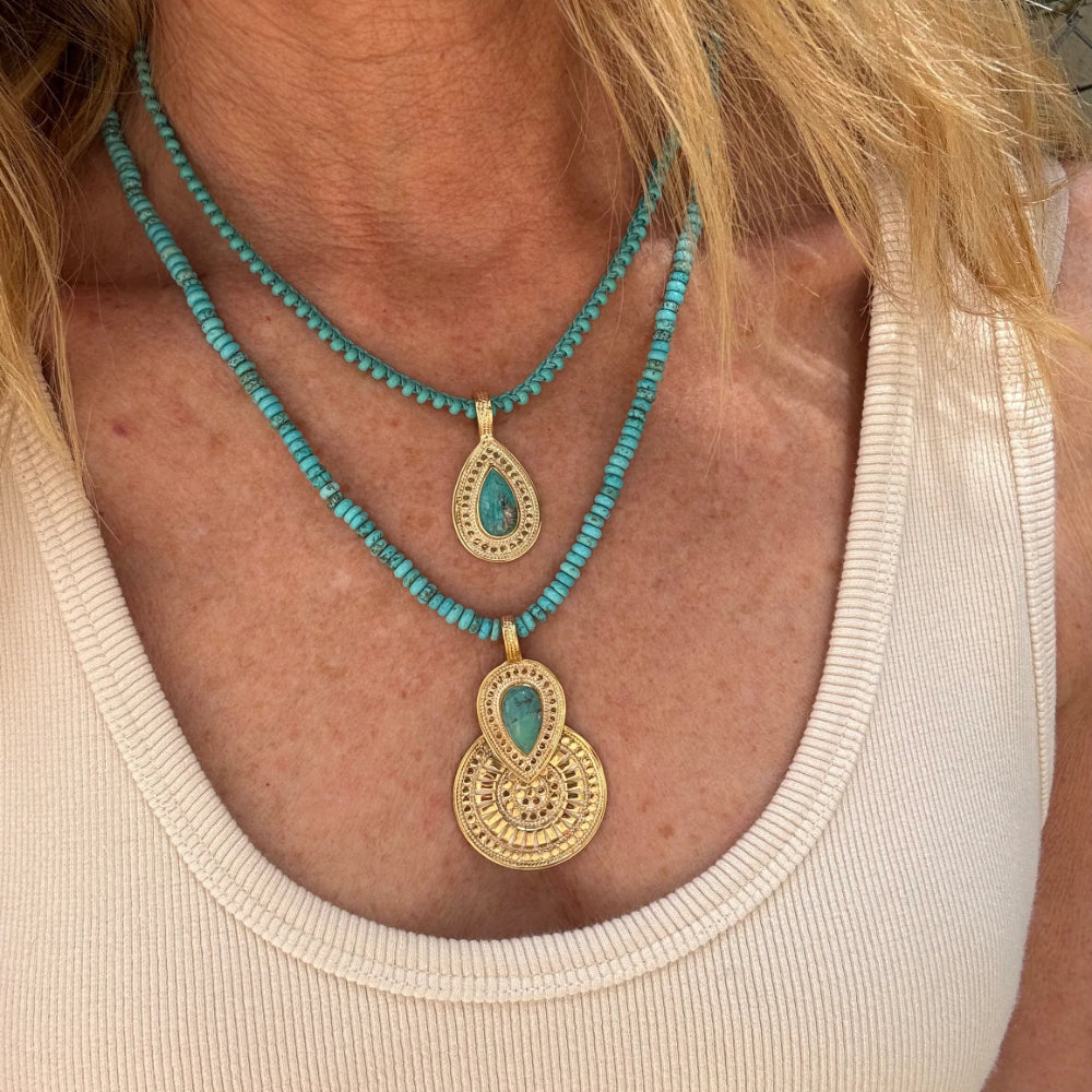 A close-up of a person wearing a layered necklace with turquoise-blue beads and gold-tone pendants, paired with a cream ribbed tank top.