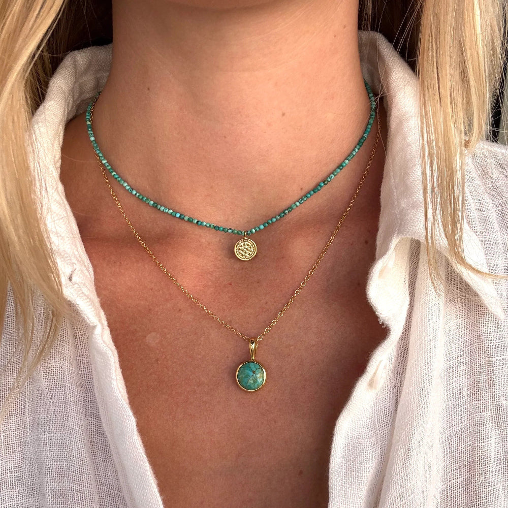 A close-up of a person's neck and chest wearing two layered necklaces: a short turquoise beaded choker with a small gold disc pendant and a longer gold chain necklace with a round turquoise pendant.