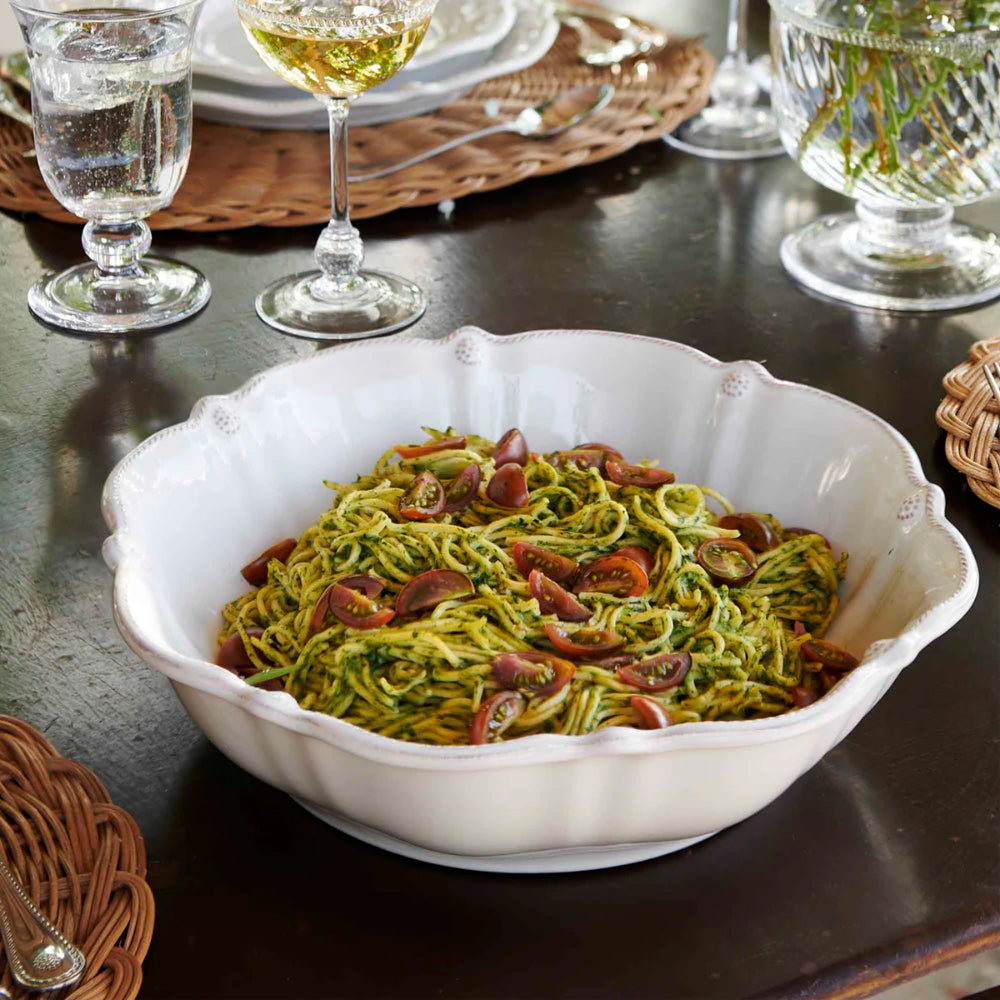 A white scalloped ceramic serving bowl filled with green pasta and sliced red olives, set on a dark wooden dining table with glasses, plates, and woven placemats in the background.