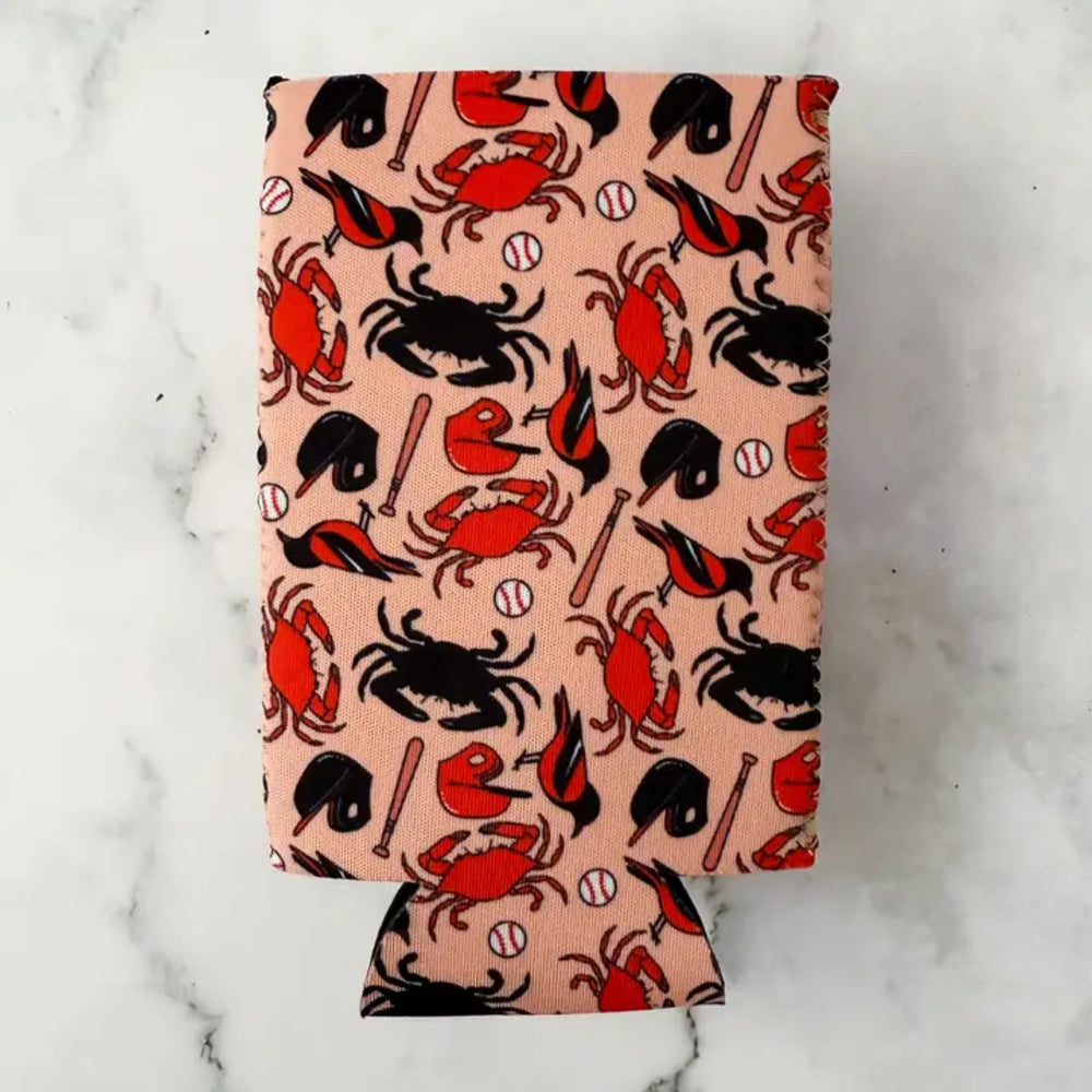 A fabric can cooler sleeve featuring a crab and baseball pattern on a peach background, photographed flat on a white marble surface.