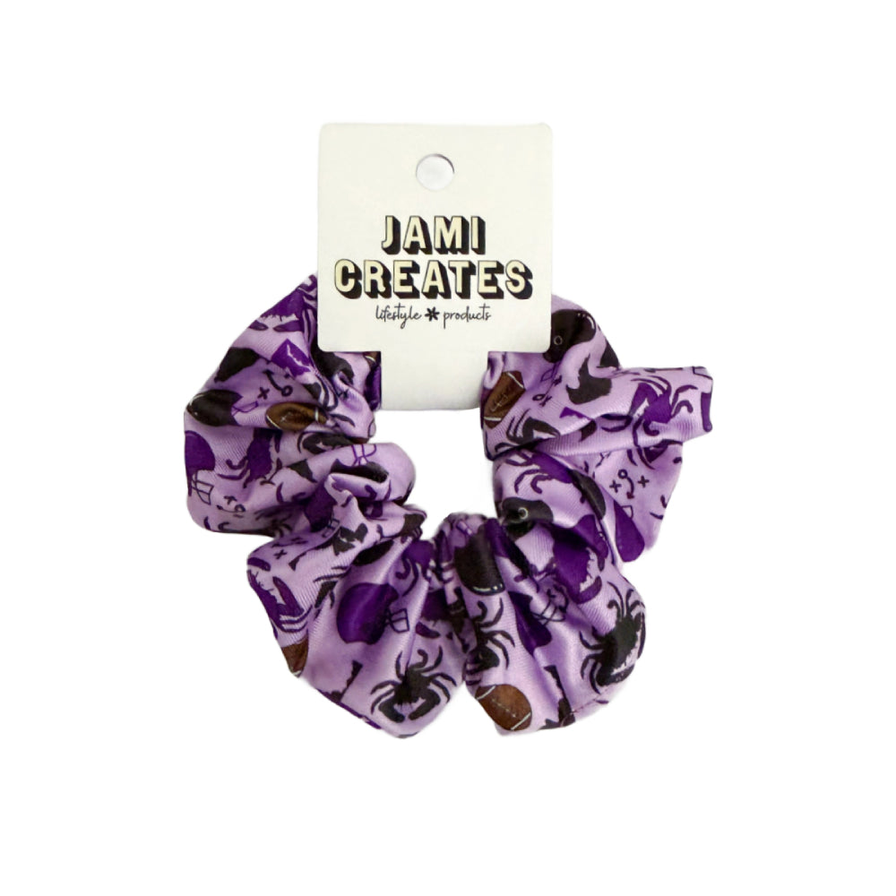 A purple fabric scrunchie with a spider and spiderweb pattern, attached to a white cardboard display card that reads 'JAMI CREATES lifestyle products' on a plain white background.