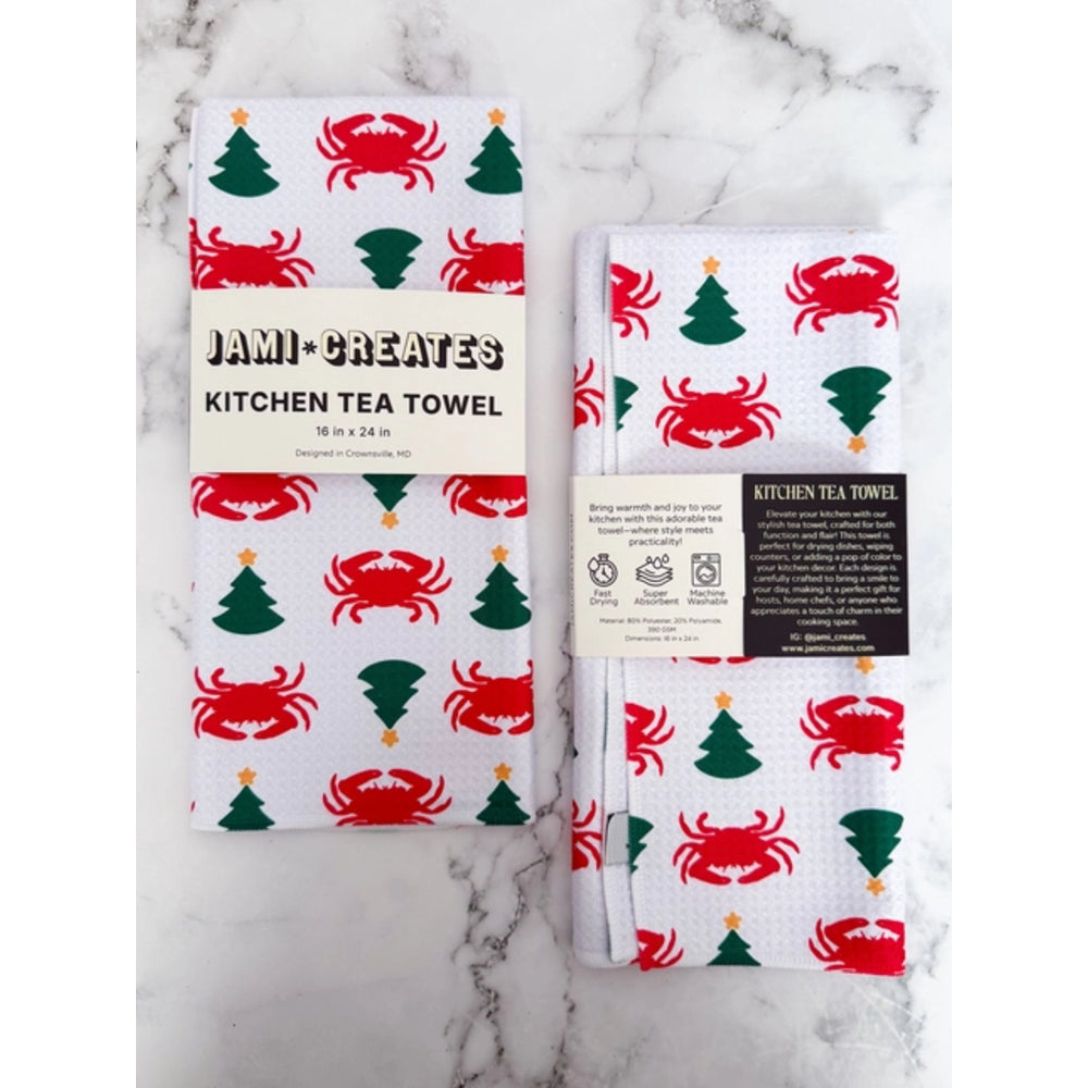 Two Jami Creates kitchen tea towels with a Christmas crab and tree pattern, displayed on a white marble background. One towel is standing upright showing the front pattern and paper band, and the other is folded showing the reverse side and paper insert.