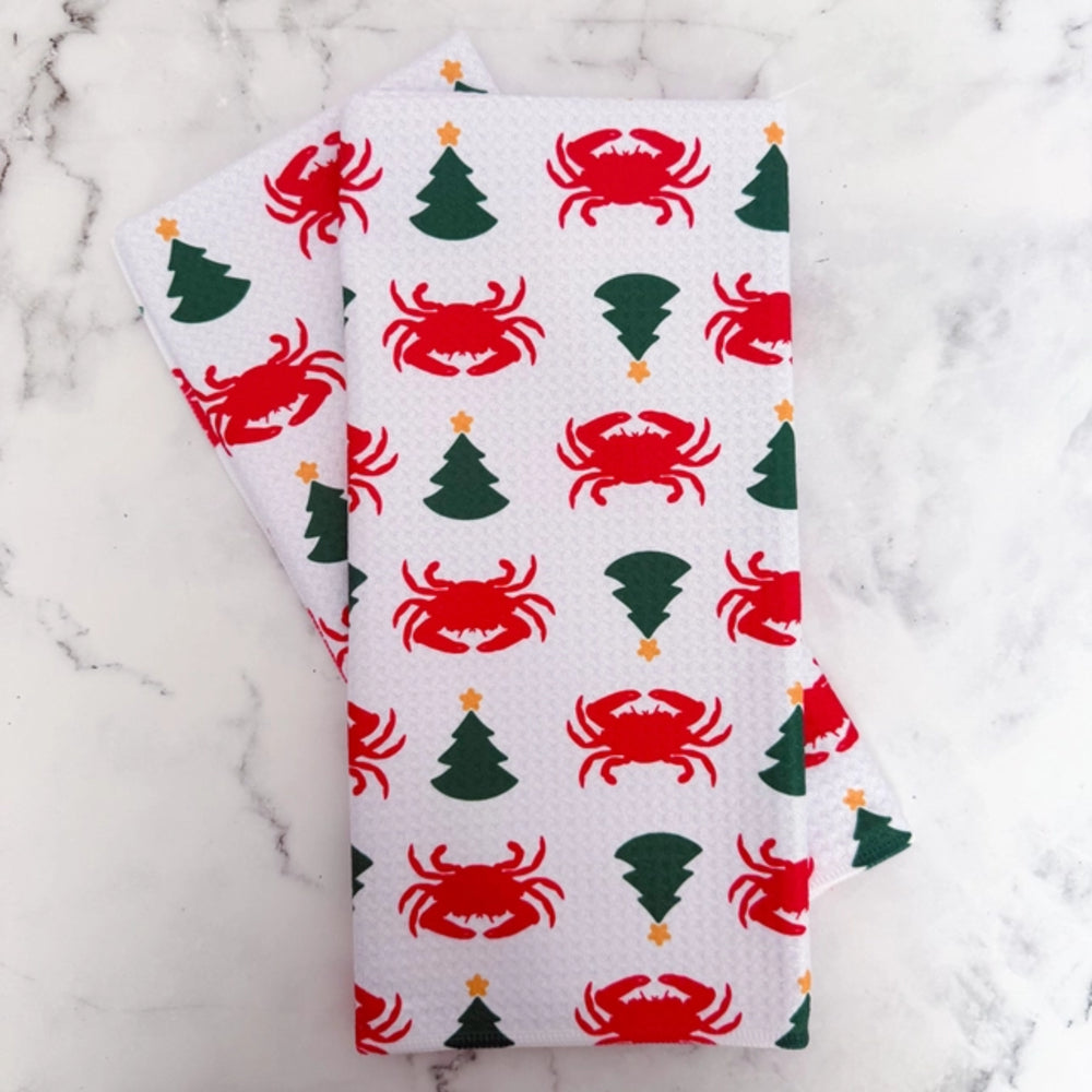 A folded white kitchen towel or napkin with a repeating holiday print of red crabs and green Christmas trees with small gold stars, displayed on a white marble surface.