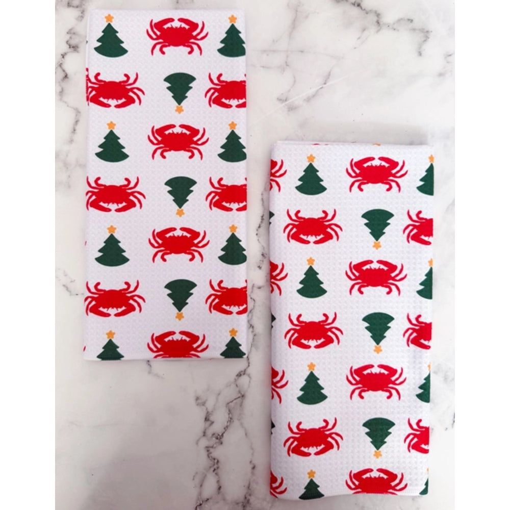 Two white kitchen towels with a festive Christmas pattern of red crabs and green Christmas trees, displayed on a white marble surface.