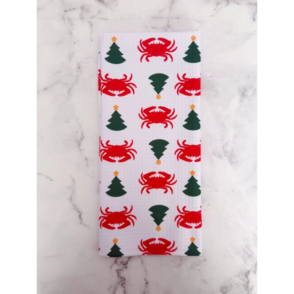 A rectangular white fabric item, likely a napkin or small towel, featuring a repeating pattern of red crabs and green Christmas trees with small gold accents, laid flat on a white marble surface.