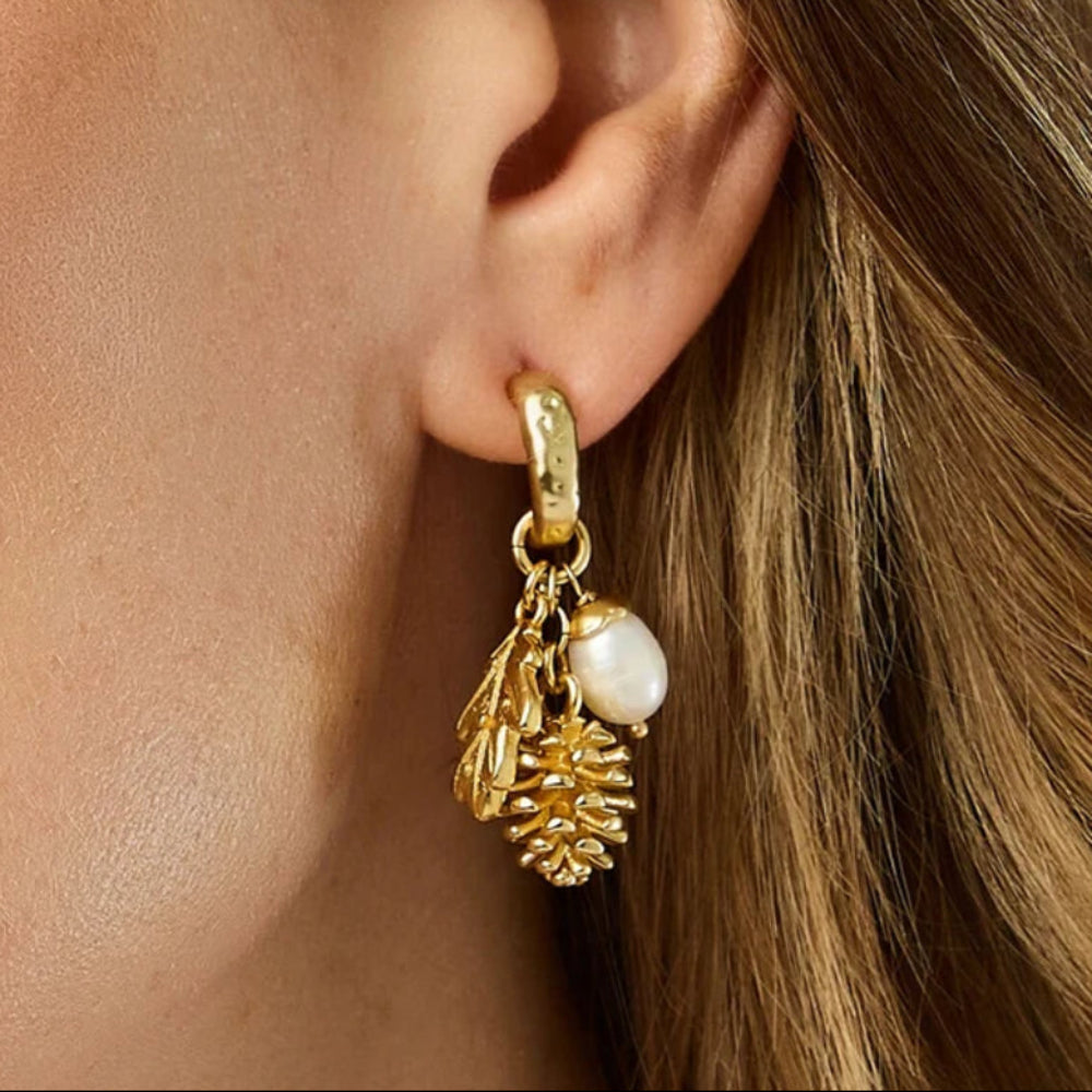 A close-up of a model's ear wearing a gold huggie hoop earring with dangling leaf and pinecone charms and a small white pearl.