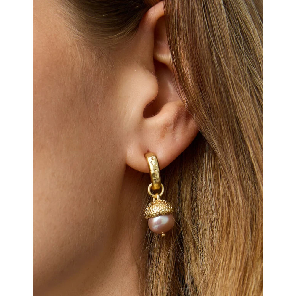 A close-up of a person's ear wearing a gold huggie hoop earring with a textured acorn-shaped charm and a dangling pearl.