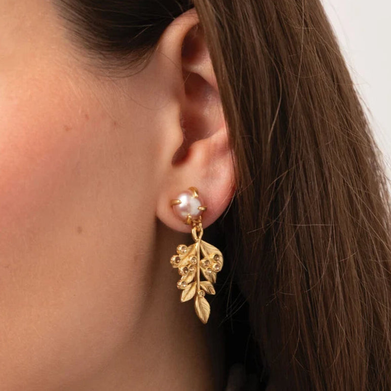 A close-up side profile of a woman wearing a delicate gold leaf dangle earring with a small white pearl stud at the top, set against a clean white background.