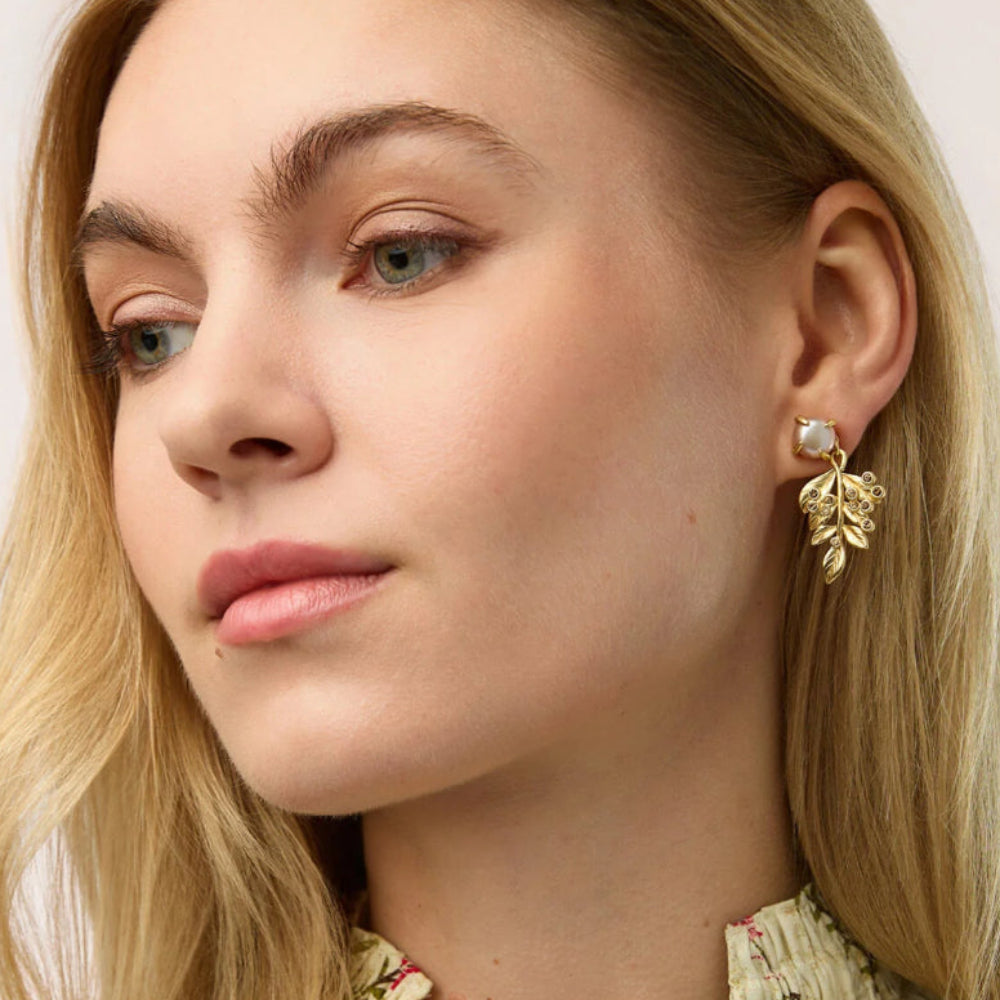 A close-up of a blonde woman wearing a gold leaf-shaped drop earring with a small white pearl accent, set against a clean off-white background.