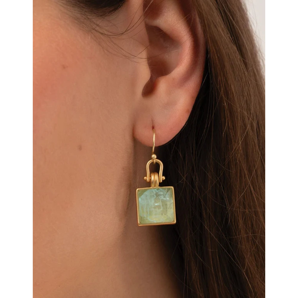 A close-up of a model's ear wearing a gold drop earring with a square light green gemstone pendant.