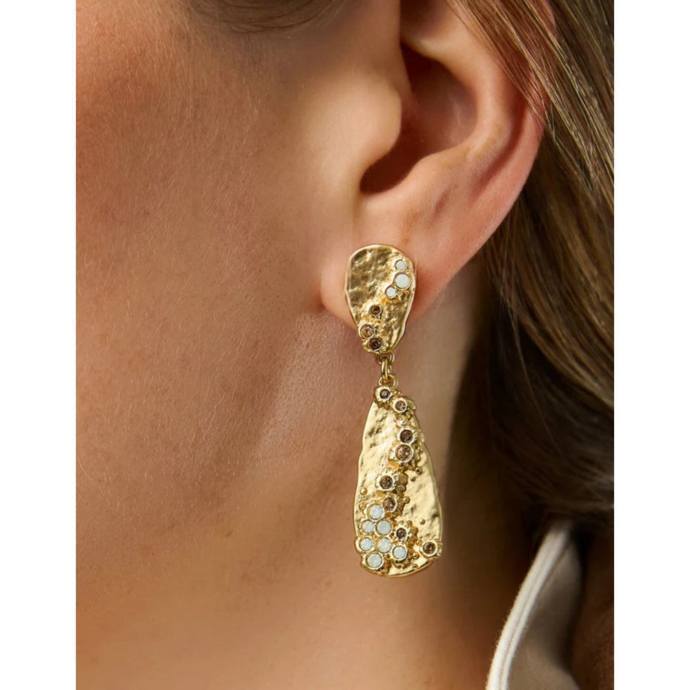 A close-up of a model's ear wearing a textured gold dangle earring adorned with small white and gold floral details.