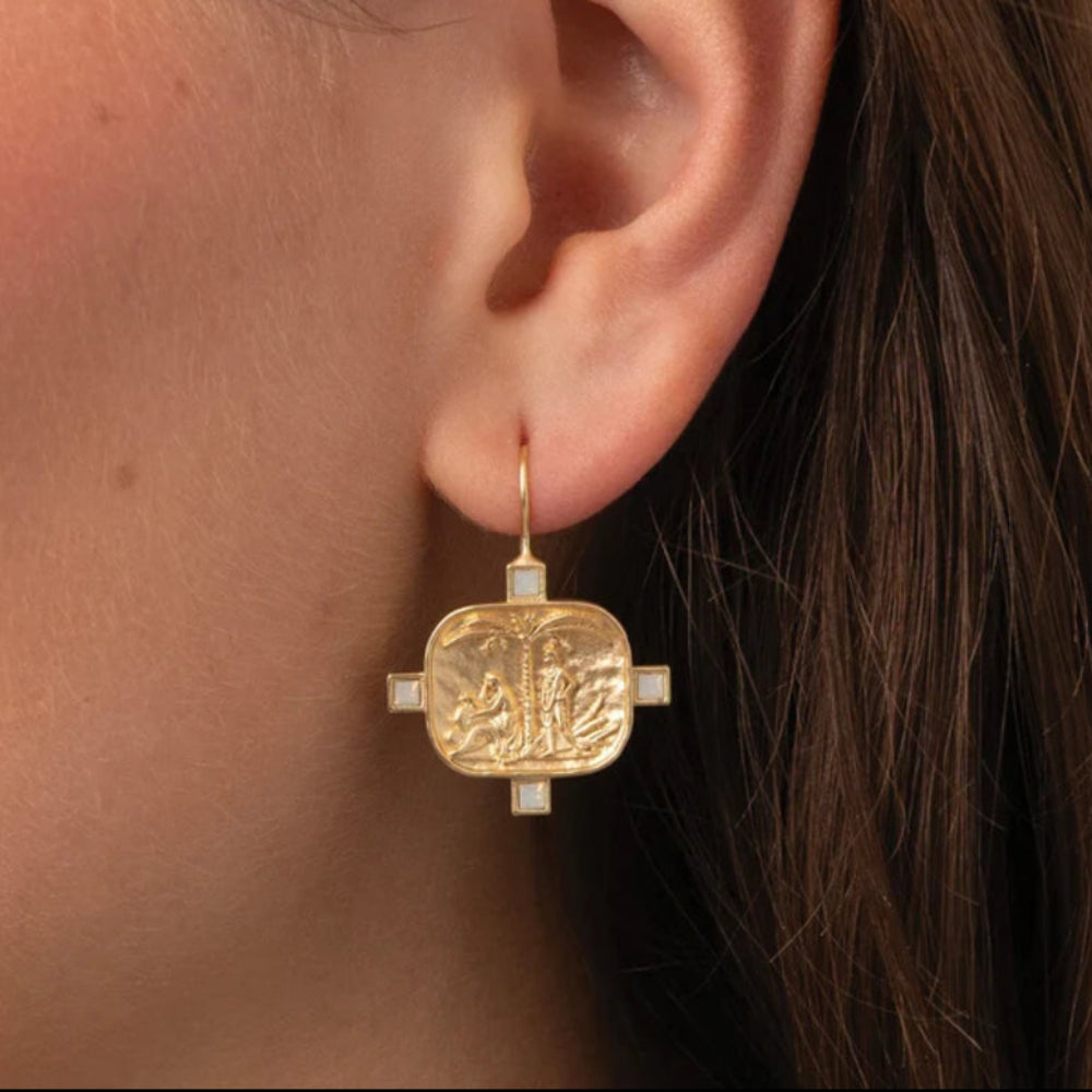 A close-up of a model's ear wearing a gold drop earring with a square, embossed design and small white accents.
