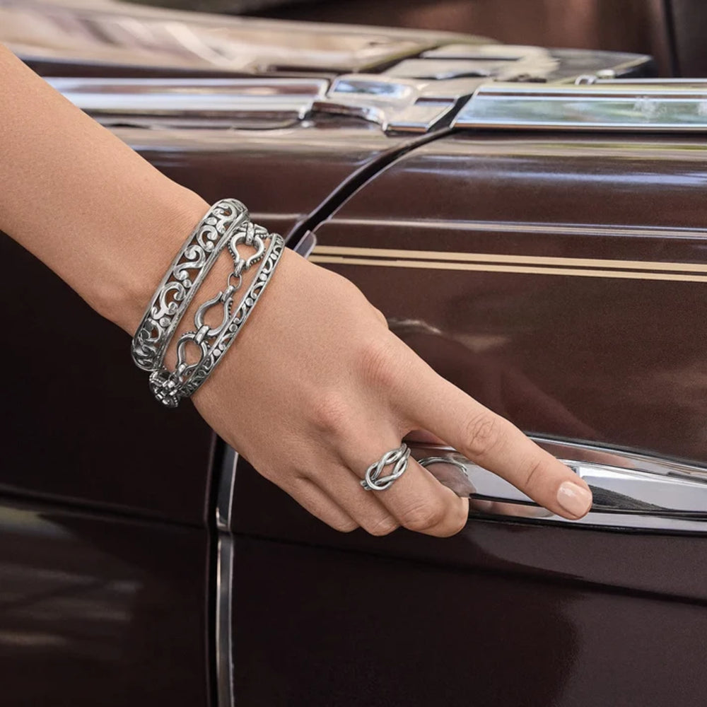A close-up of a person's hand and wrist wearing a stack of three ornate silver chain-link bracelets, reaching for a chrome handle on a vintage dark brown car.