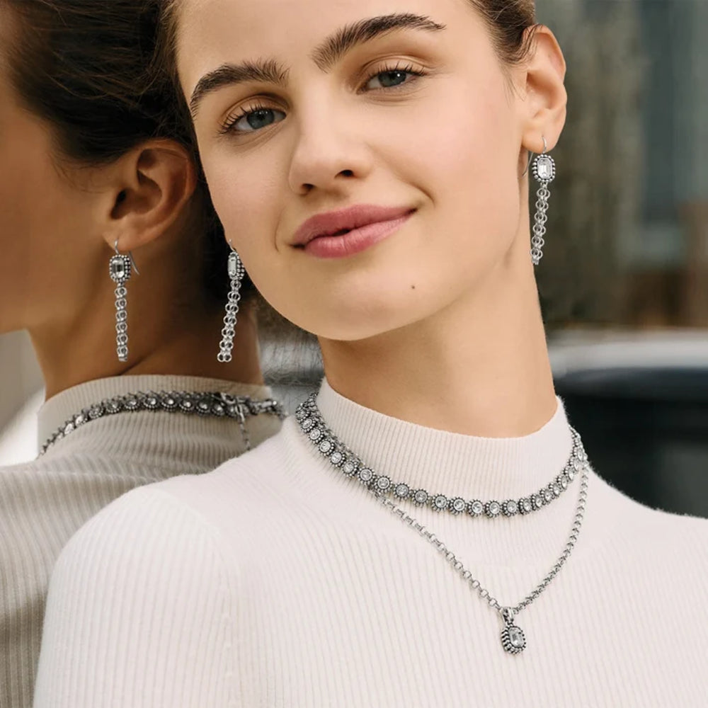 Woman wearing silver jewelry including earrings and necklaces, with a blurred background.