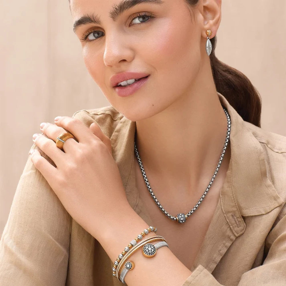 A close-up lifestyle shot of a woman wearing a silver beaded necklace with a central floral starburst, matching silver and gold bangles, and a gold ring, styled with a beige linen shirt against a soft beige background.