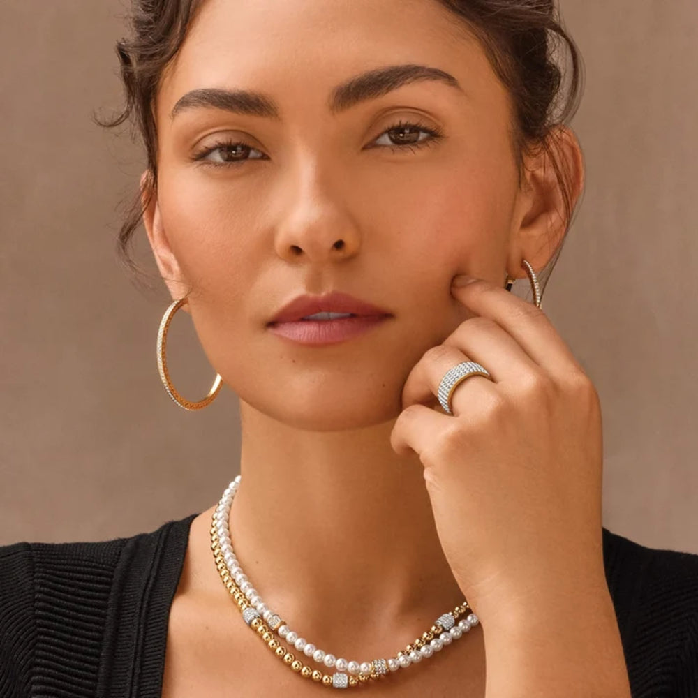 A close-up of a woman wearing a double-strand pearl and gold beaded necklace, matching gold hoop earrings, and a textured gold ring.