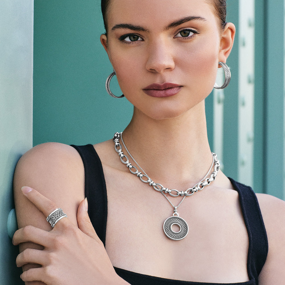 Woman wearing silver jewelry including a necklace, hoop earrings, and ring against a teal background