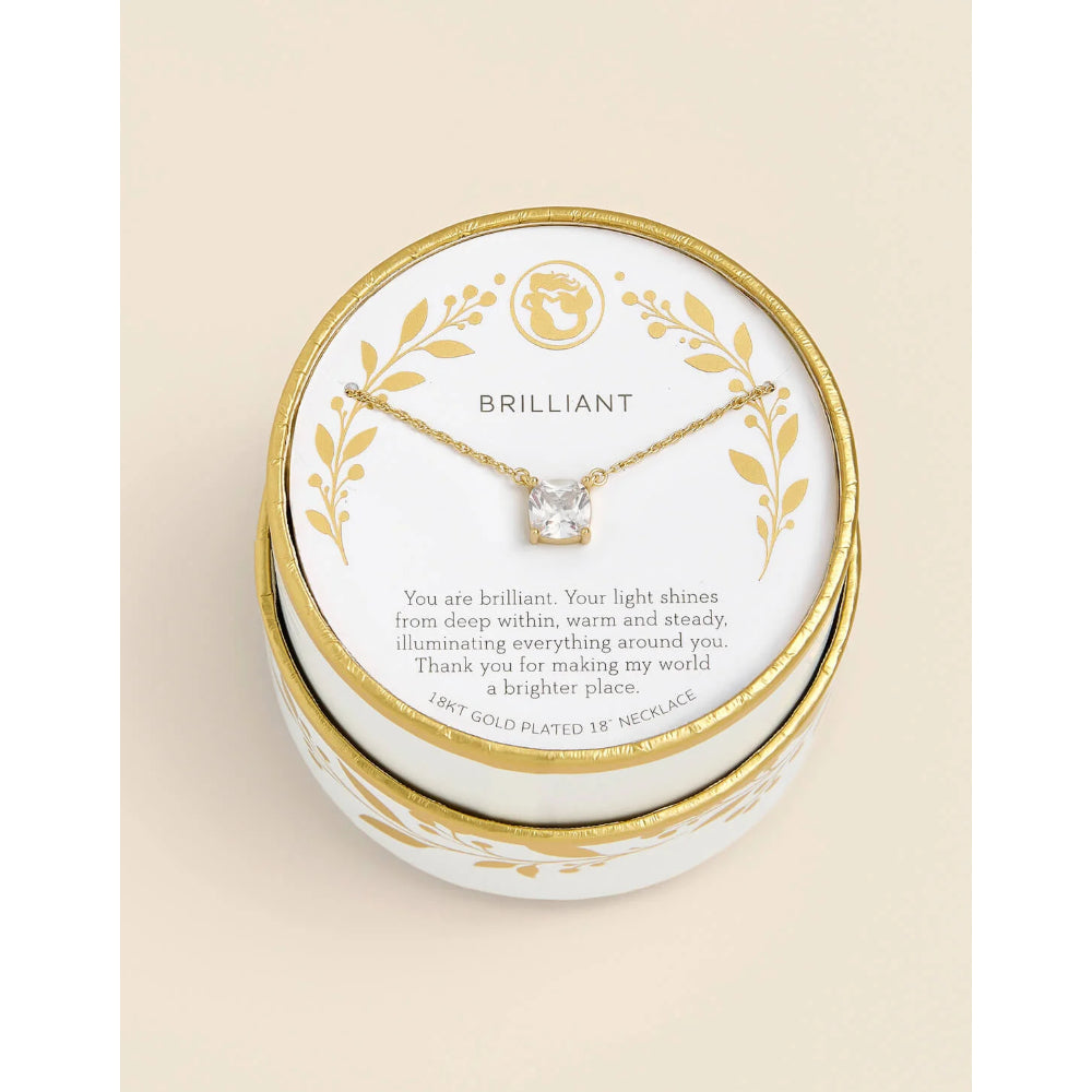 A gold-plated 18k necklace with a clear gemstone pendant, presented on a white card with gold leaf illustrations and the brand name BRILLIANT, inside a round white and gold gift box.