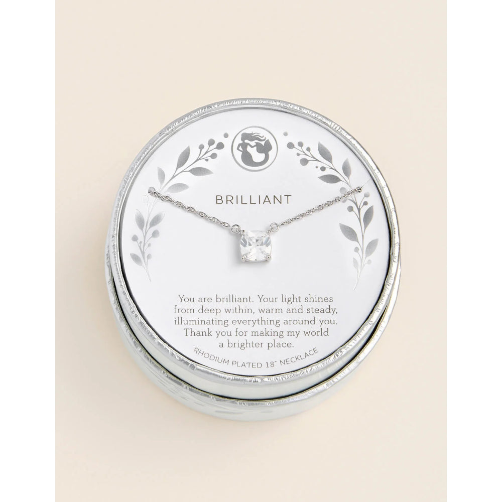 A silver-plated necklace with a clear gemstone displayed on a round white card inside a silver presentation box. The card is engraved with floral designs and the word 'BRILLIANT' above a touching quote.