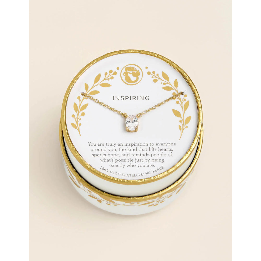 A gold-plated 18-mille necklace with a clear gemstone pendant, displayed on a white and gold round card inside a matching round presentation box. The card features the word 'INSPIRING' and a quote, set against a warm cream background.
