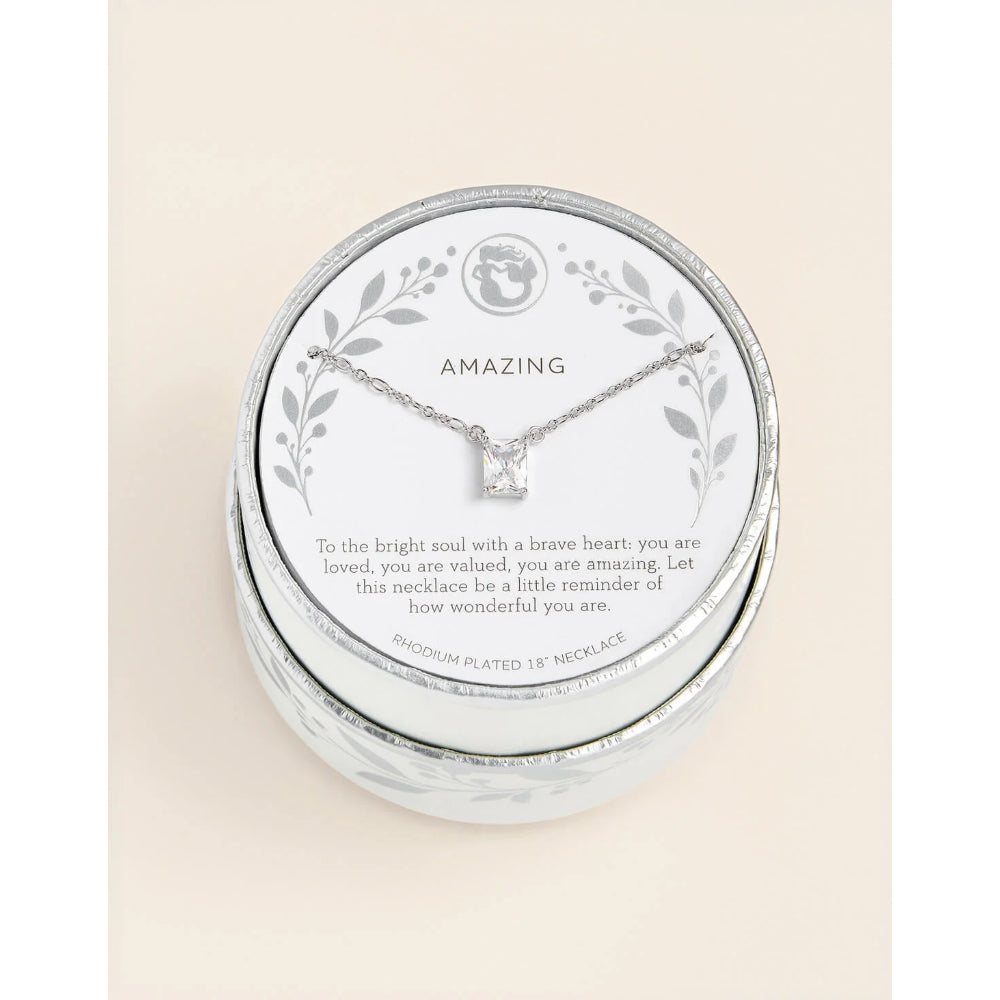 A silver necklace with a square-cut clear gemstone displayed on a branded round presentation card inside a silver tin box. The card reads 'AMAZING' and includes a sentimental message, photographed against a light cream background.