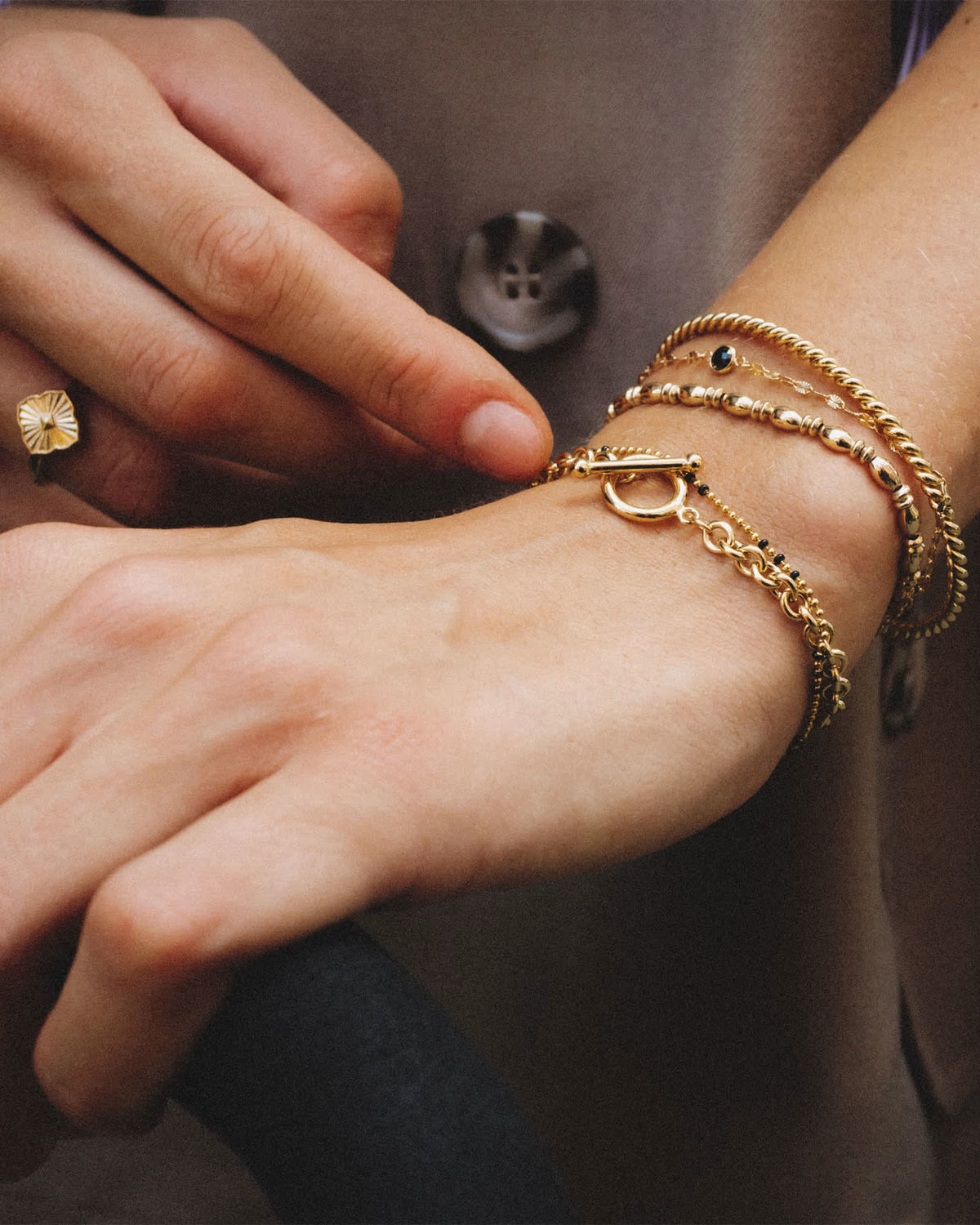 A close-up of a person's wrist wearing multiple delicate gold bracelets, including a rope-style bracelet, a beaded chain bracelet, and a thin chain bracelet with a toggle clasp. A matching gold ring is also visible on the finger.