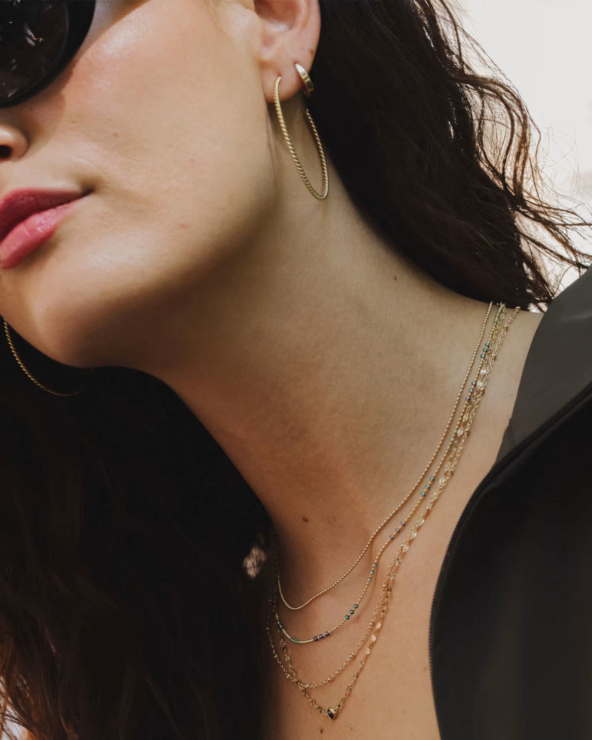 Close-up of a woman wearing layered gold necklaces, a gold hoop earring, and sunglasses, with dark hair and a black top visible.