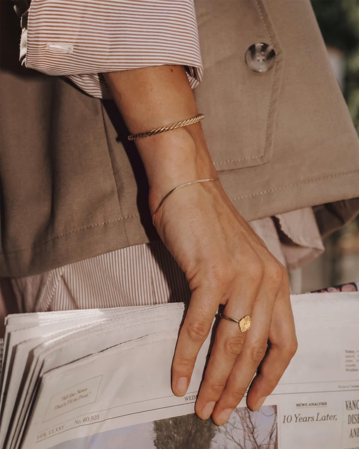 A close-up lifestyle shot of a person's hand wearing a gold ring with a textured square face and a thin gold bangle, while holding a stack of papers.