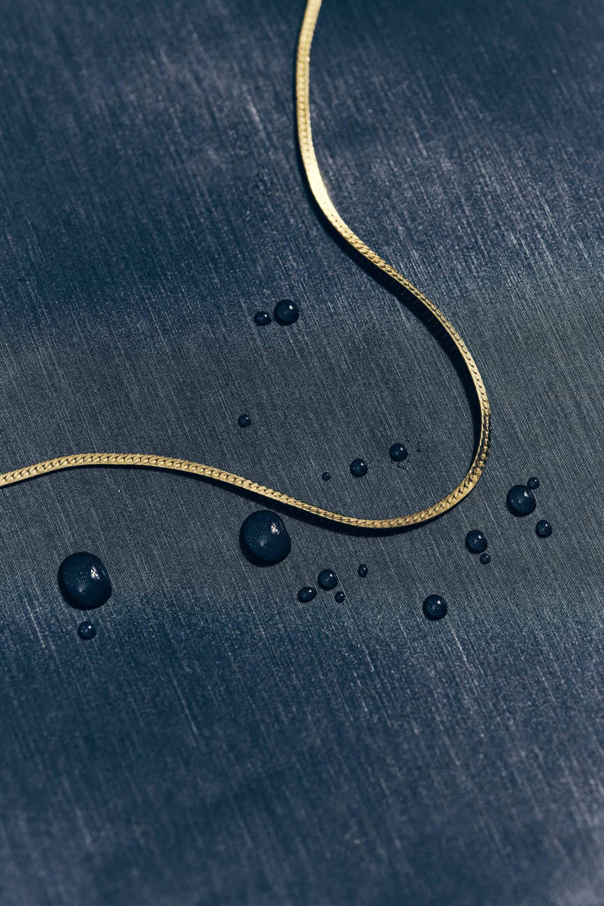 A close-up of a thin gold chain laid on a dark blue textured surface with water droplets scattered around it.