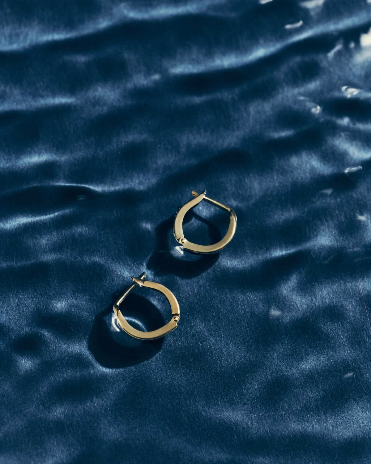 A pair of gold hoop earrings resting on a dark blue textured surface with soft ripples.