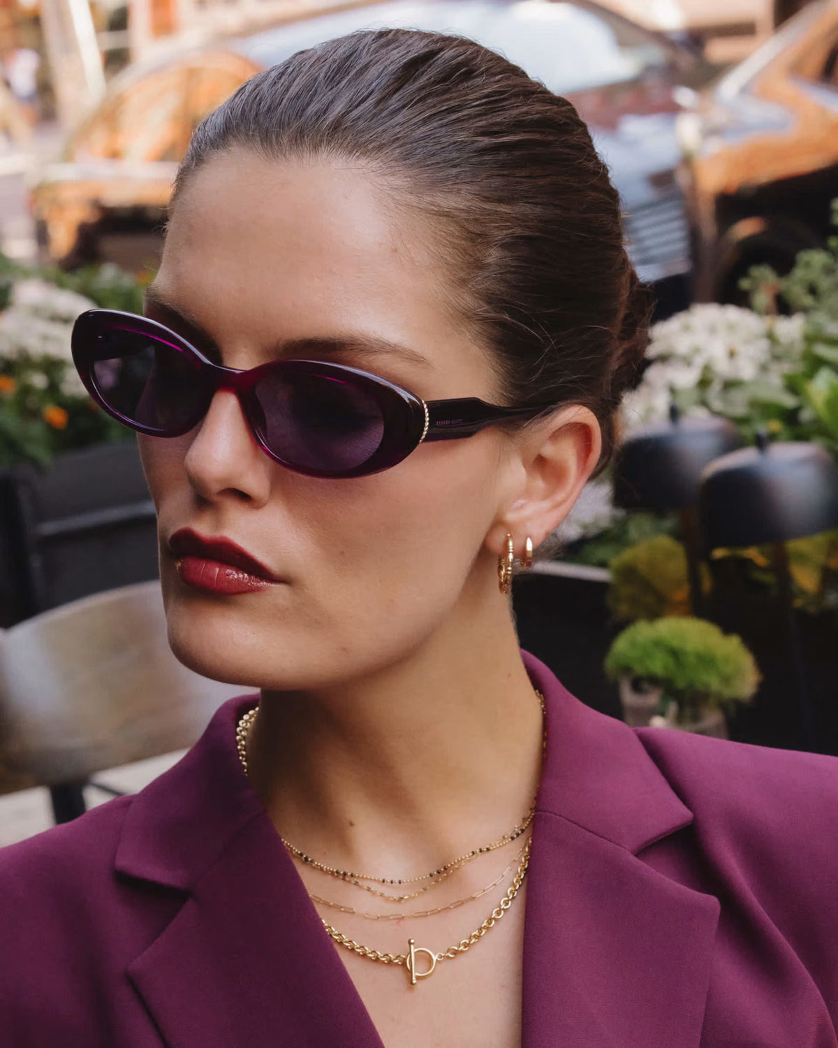 A close-up lifestyle shot of a woman wearing dark purple cat-eye sunglasses, a magenta blazer, and gold layered necklaces, set against a blurred outdoor background.